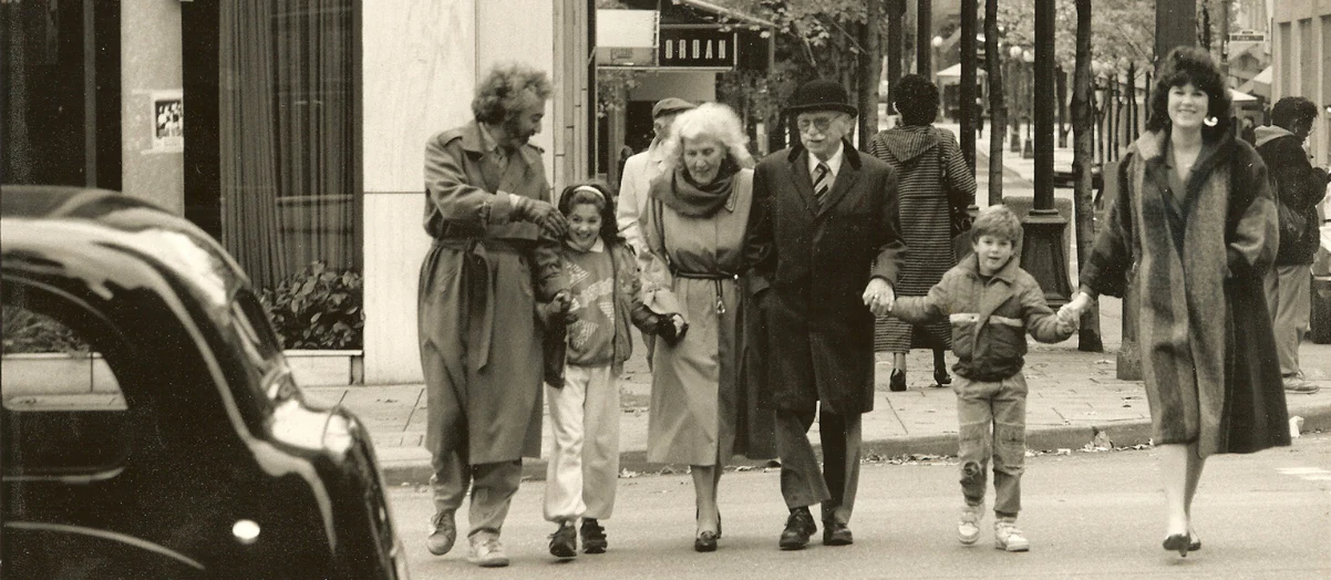 Black and white photo of a family of 6 people walking across a street with a car in the foreground 