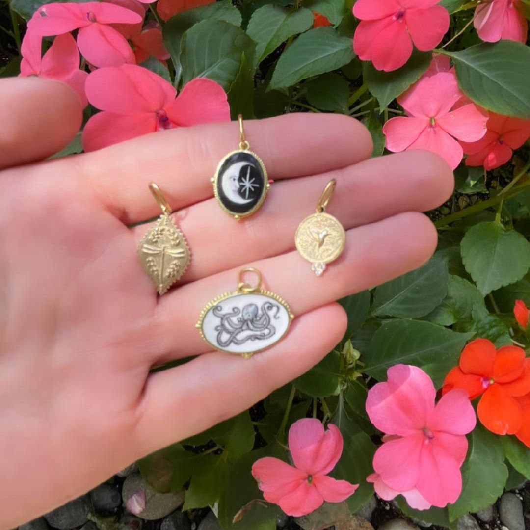 Erica Molinari charms in the palm of a woman's hand. The charms show the hand painted moon enamel and octopus, as well as the dragonfly and hummingbird charms in yellow gold