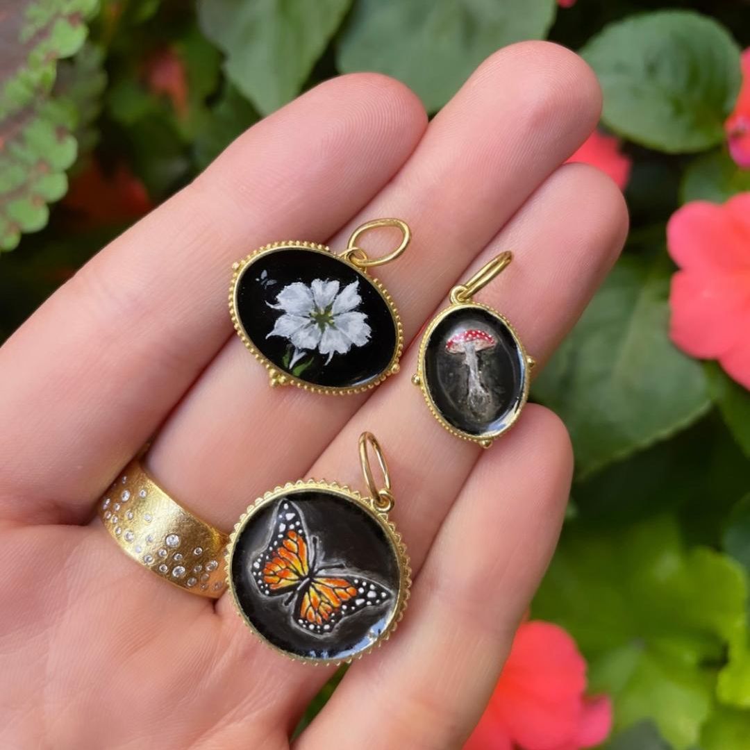 3 Hand painted charms in yellow gold on a hand. One white flower, one monarch butterfly and one mushroom.