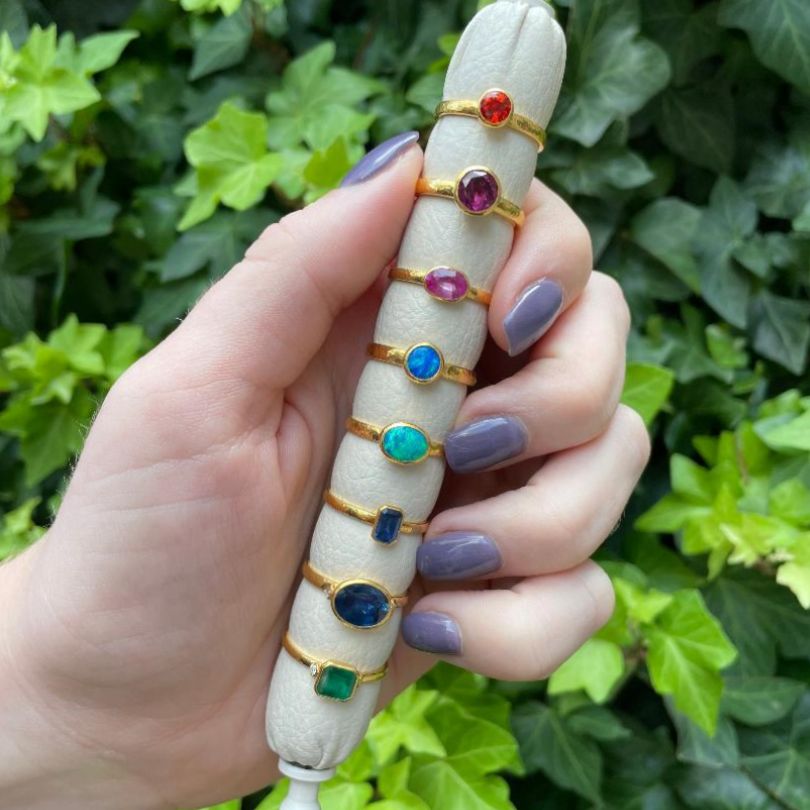 rainbow of colored stone rings in 18K yellow gold being held with a green background