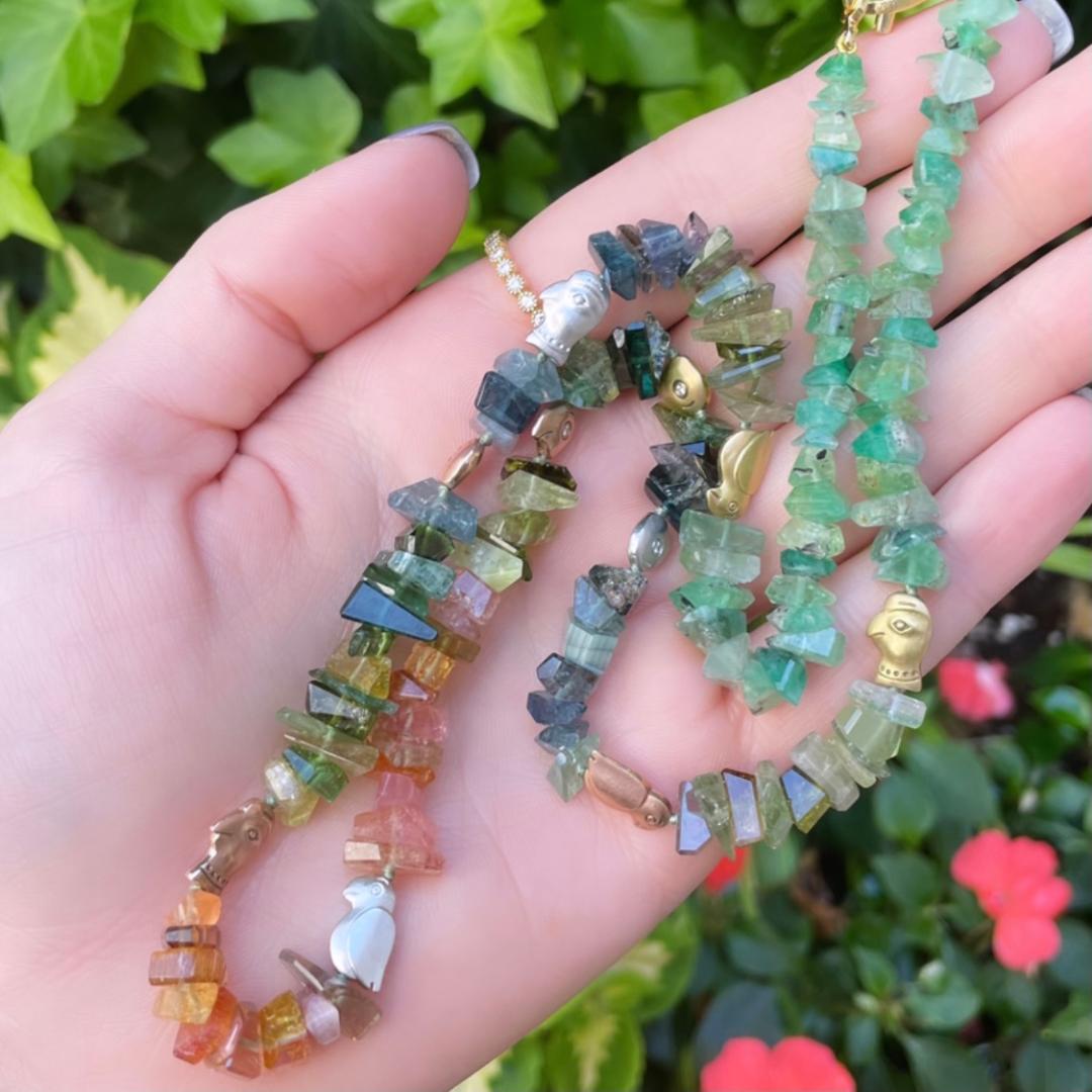 multicolored tourmaline necklace with gold and palladium characters held on a palm over a green background