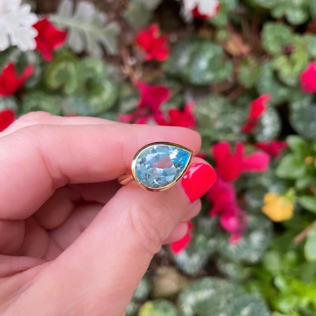 Hand holding a blue pear shape yellow gold ring in front of flowers