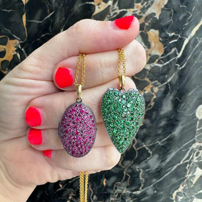Silver lockets in red ruby and green tsavorite being held in a hand over a black marble background