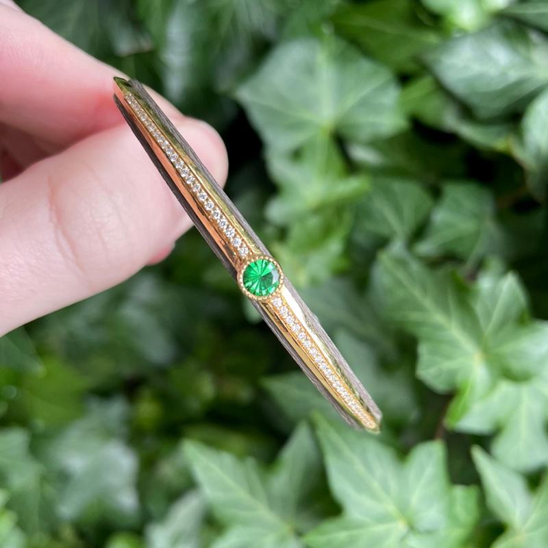 Fingers holding a silver and gold bangle with a green stone and diamonds
