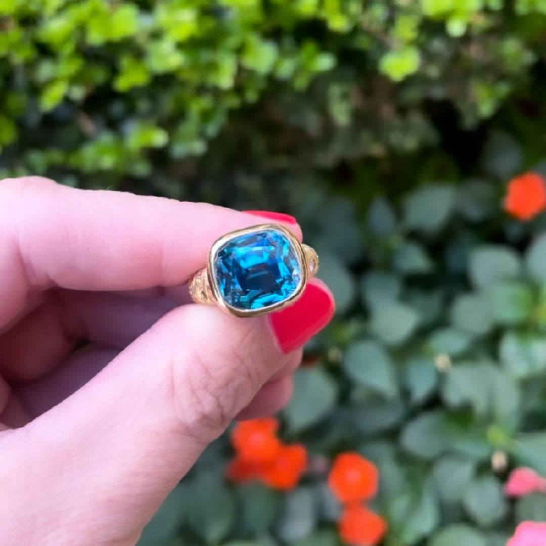 Blue gemstone and gold ring being held by fingers in front of a bush
