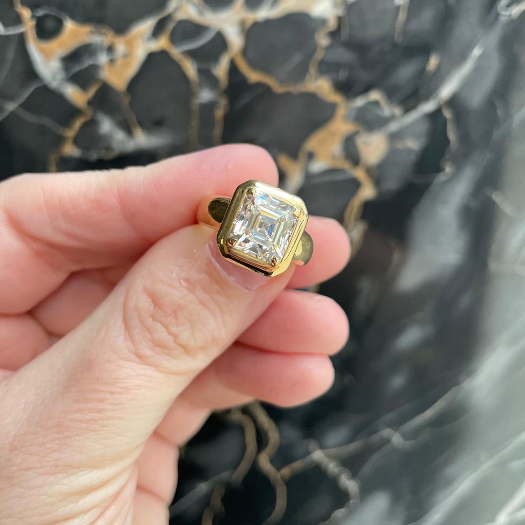 Square diamond ring in yellow gold being held against a dark marble background