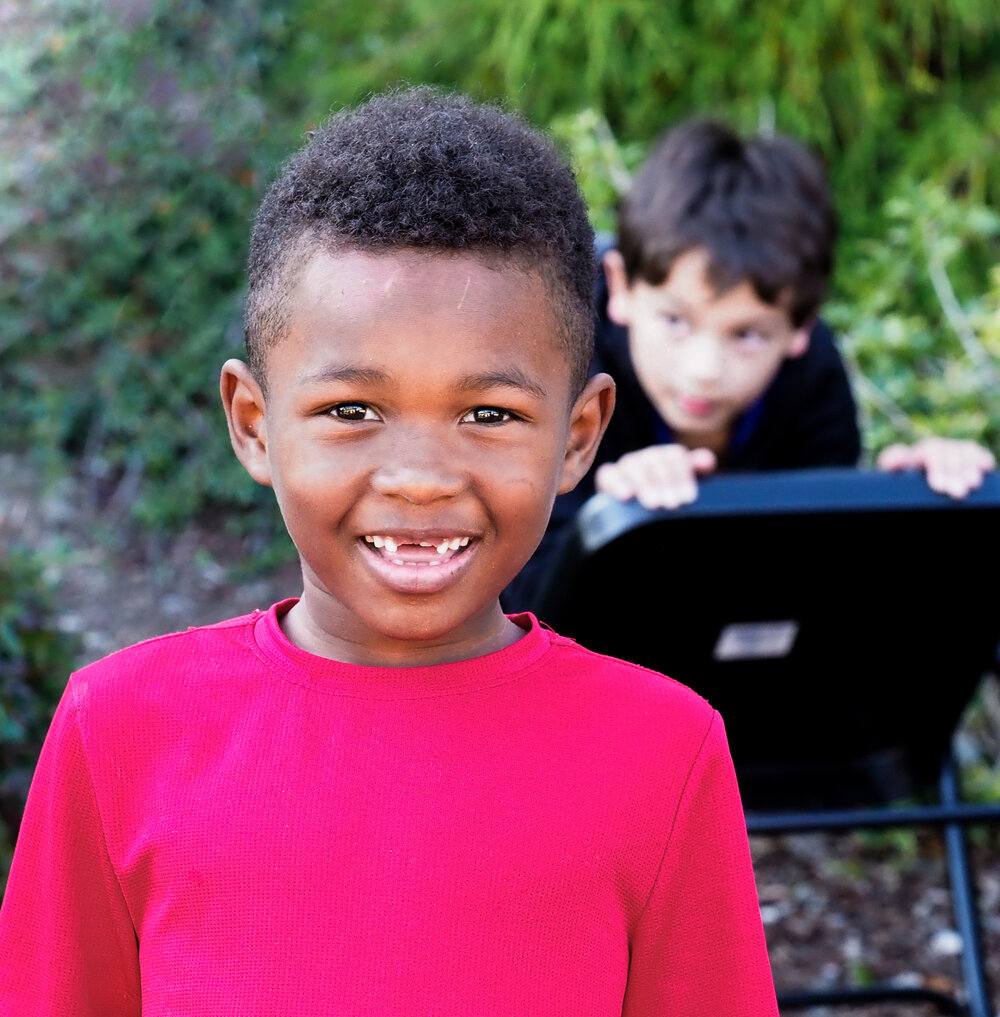 little boy missing his front teeth looking into camera and smiling with another child in the back ground.
