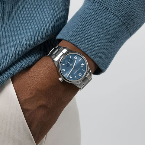 close up on persons wrist with hand in pocket wearing a blue sweater, white pants and stainless steel blue dial watch on wrist