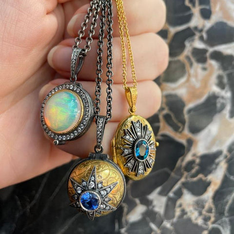 gold and silver lockets with opal, sapphire, and tourmaline center stones being held in a hand with a marble background