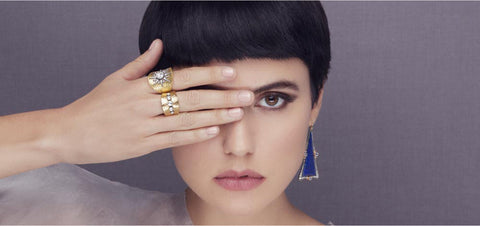 Close up of woman's face with hand covering one eye. That had has two 22k and white diamond rings on it. One blue triangular earring is shown.