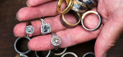 Man holding gold and silver rings in hand and three diamond rings on fingers with more rings below.