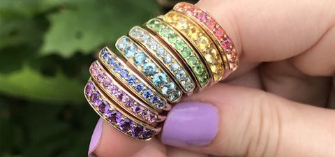 Rainbow order of Jane Taylor rings on a finger with leaves in the background.